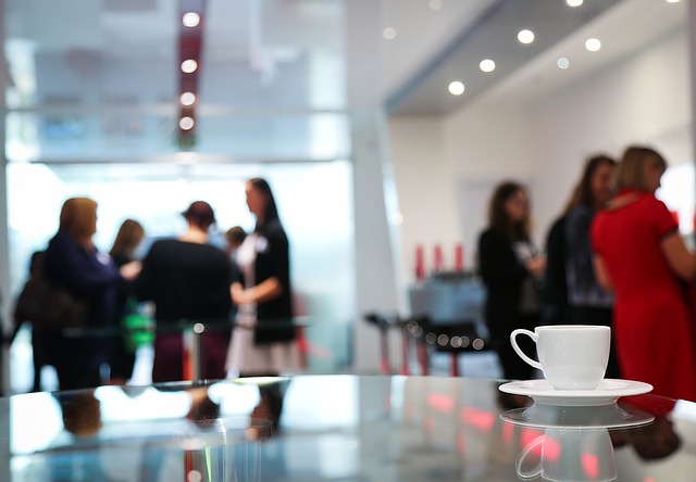 A white coffee cup sits on a table in focus while two groups of women in business attire are out of focus in the background.